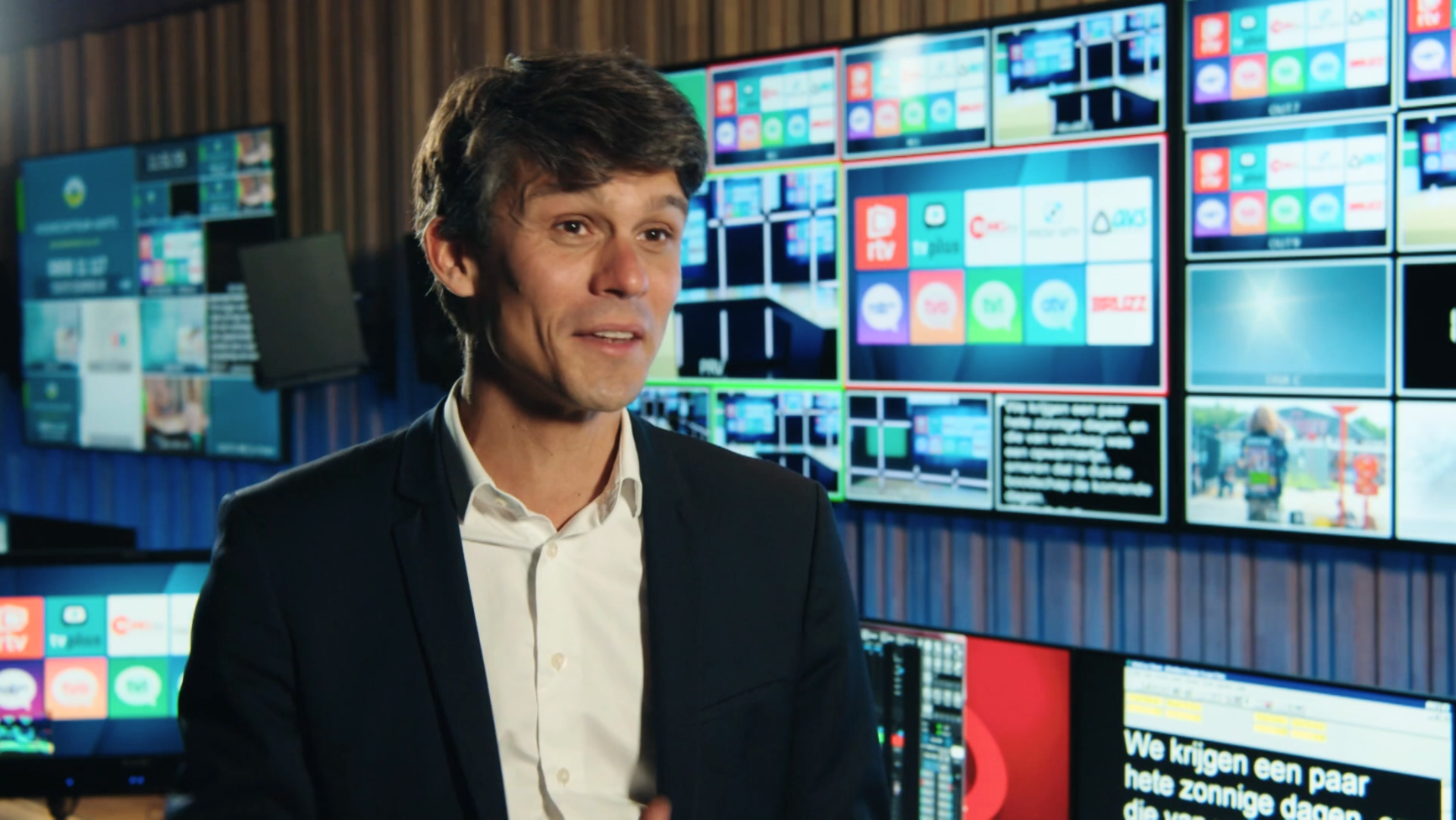 Man speaking in front of multiple screen displays showing broadcast interfaces and content.